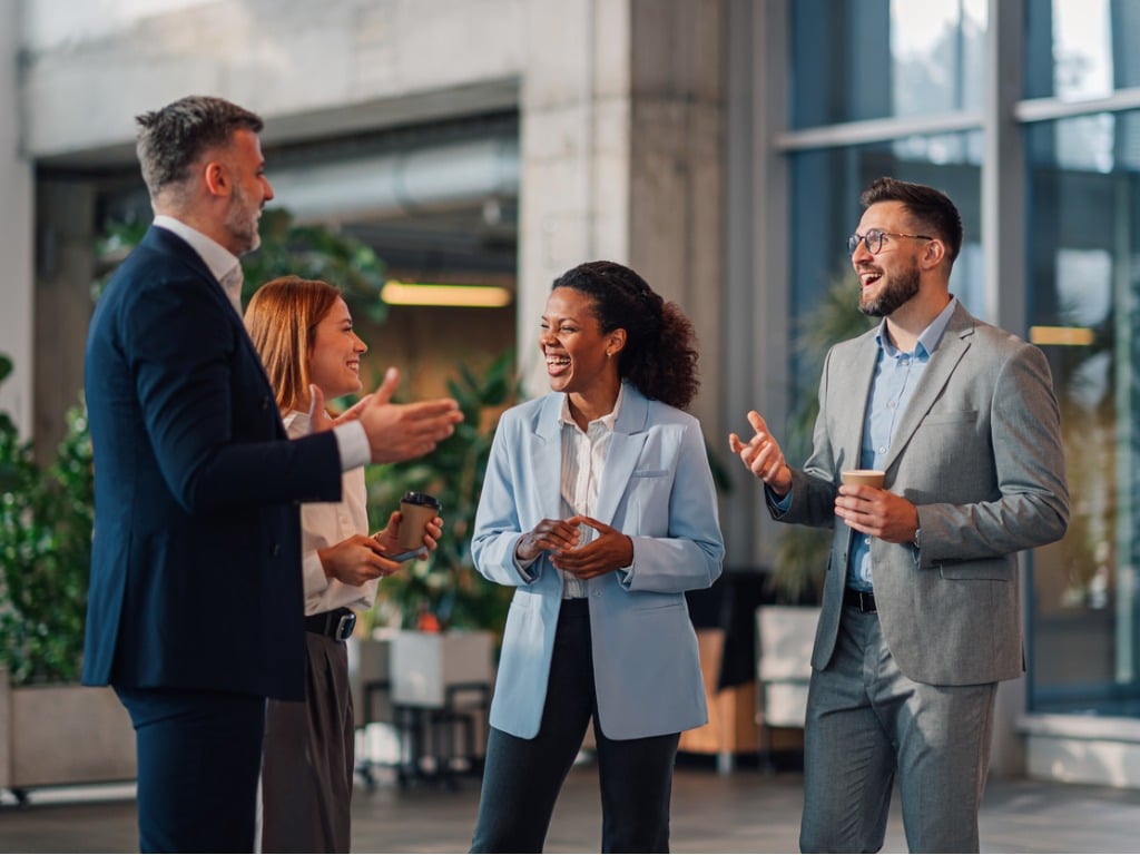 Four diverse business professionals in conversation, smiling and gesturing in a modern office setting.