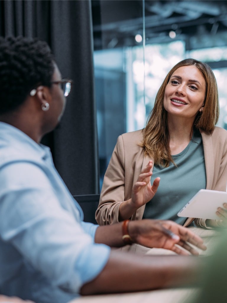 Two women in business attire discuss something at a table with a tablet.