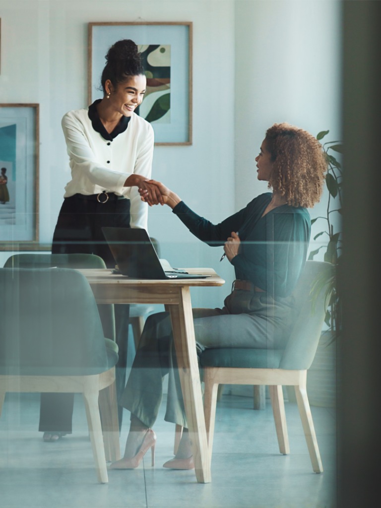 Two women shaking hands across a table in a modern office setting, smiling.